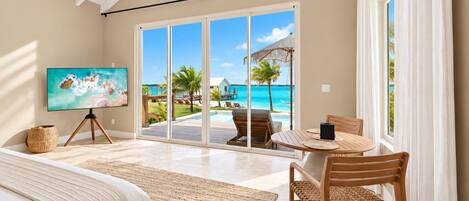 A bedroom with a large bed, a TV on a tripod, a dining table with chairs, and a view of a beach and pool through large sliding doors.