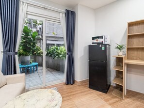A compact living space with a sliding glass door leading to a balcony, a black refrigerator, a wooden shelf unit, and a marble-patterned area rug. - YOUR HOME - Garden Studio 35m² (Ho Chi Minh City)
