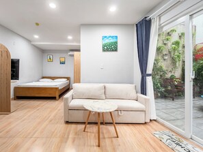 A modern bedroom with a bed, a sofa, and a small table. There is a window with curtains and a view of a garden. - YOUR HOME - Garden Studio 35m² (Ho Chi Minh City)