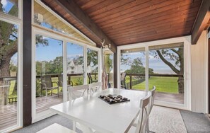 An open-plan room with a large table and chairs, a wooden ceiling, and large windows offering a view of a grassy area and trees. - Beachside Dream · Spacious 4-bedroom home in Stevensville (Stevensville)
