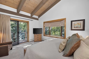A bedroom with a large bed, a TV, a dresser, and two windows offering views of trees. - Spring Hill Hideaway Fireplace (Vail)