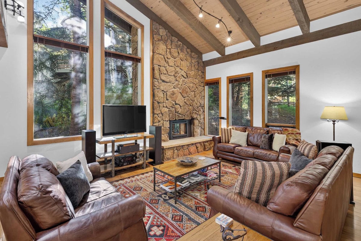 A spacious living room with a stone wall, large windows, a fireplace, leather sofas, and a flat-screen TV.