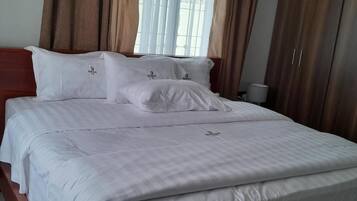 Una cama bien hecha con ropa blanca y almohadas, un cabecero de madera y una ventana con cortinas.