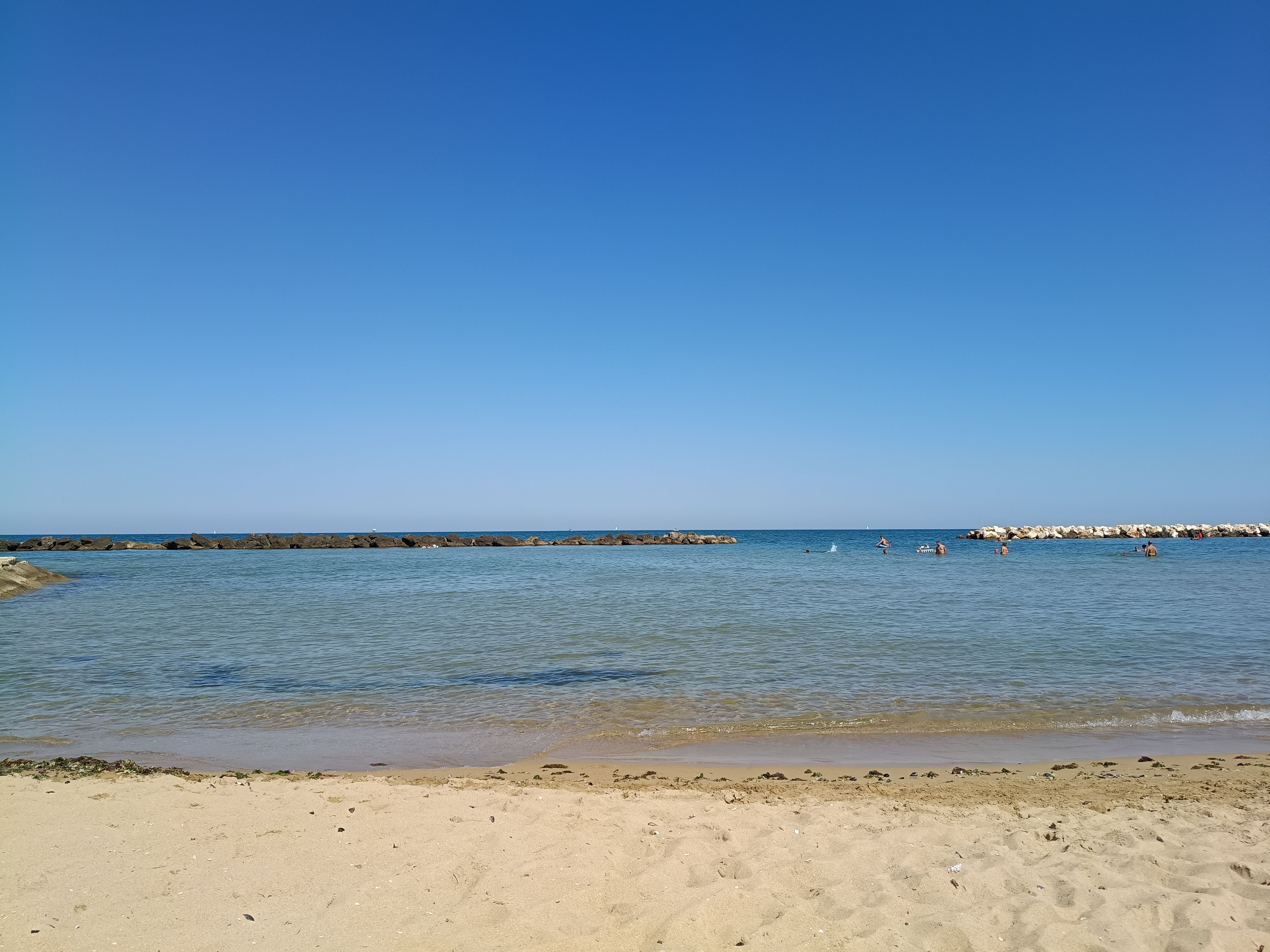 Een strand met helderblauw water, een zandstrand en een rotsachtige zeewering in de verte.