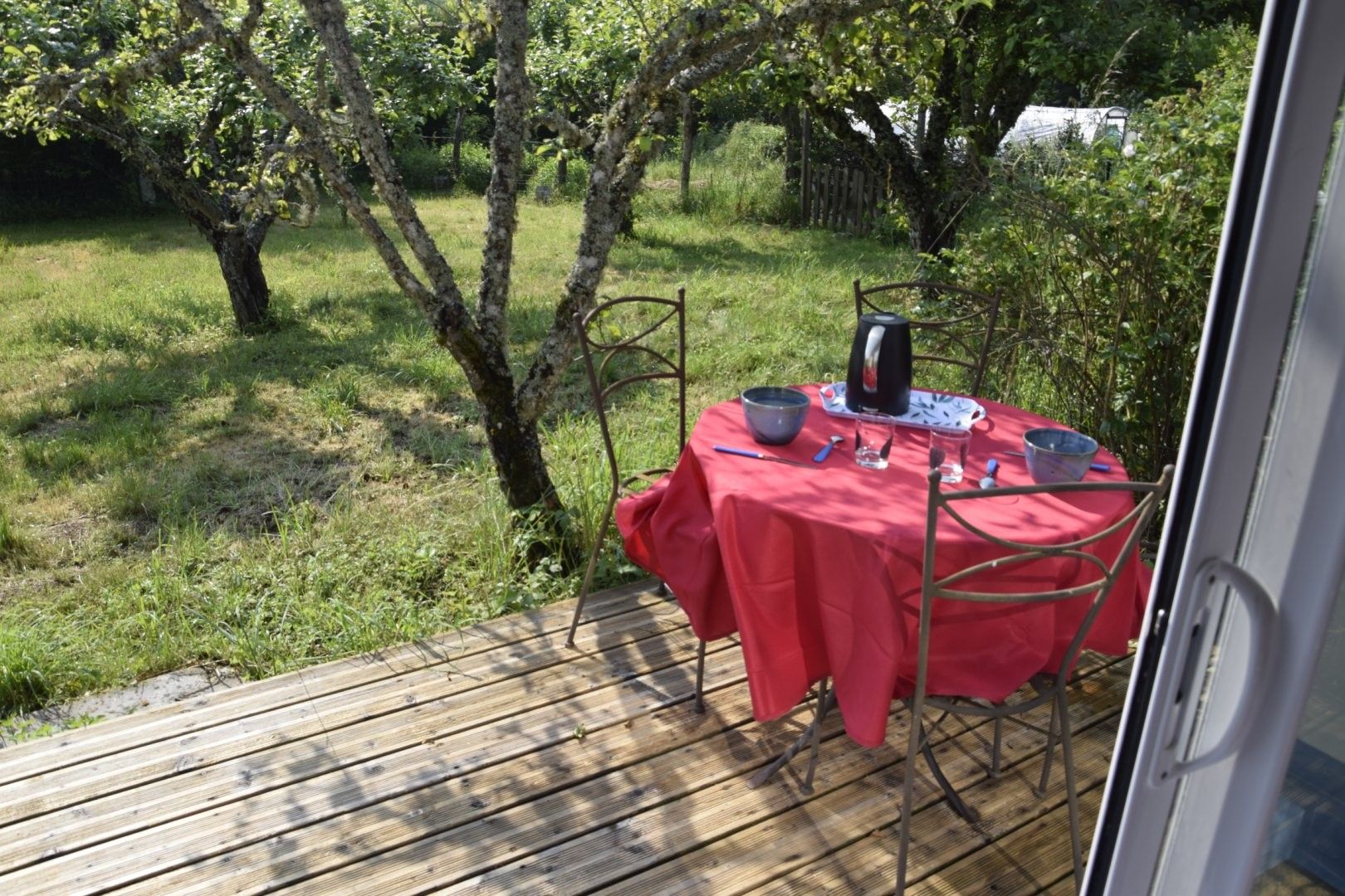 Een houten terras met een rode eettafel, omgeven door groen en bomen.