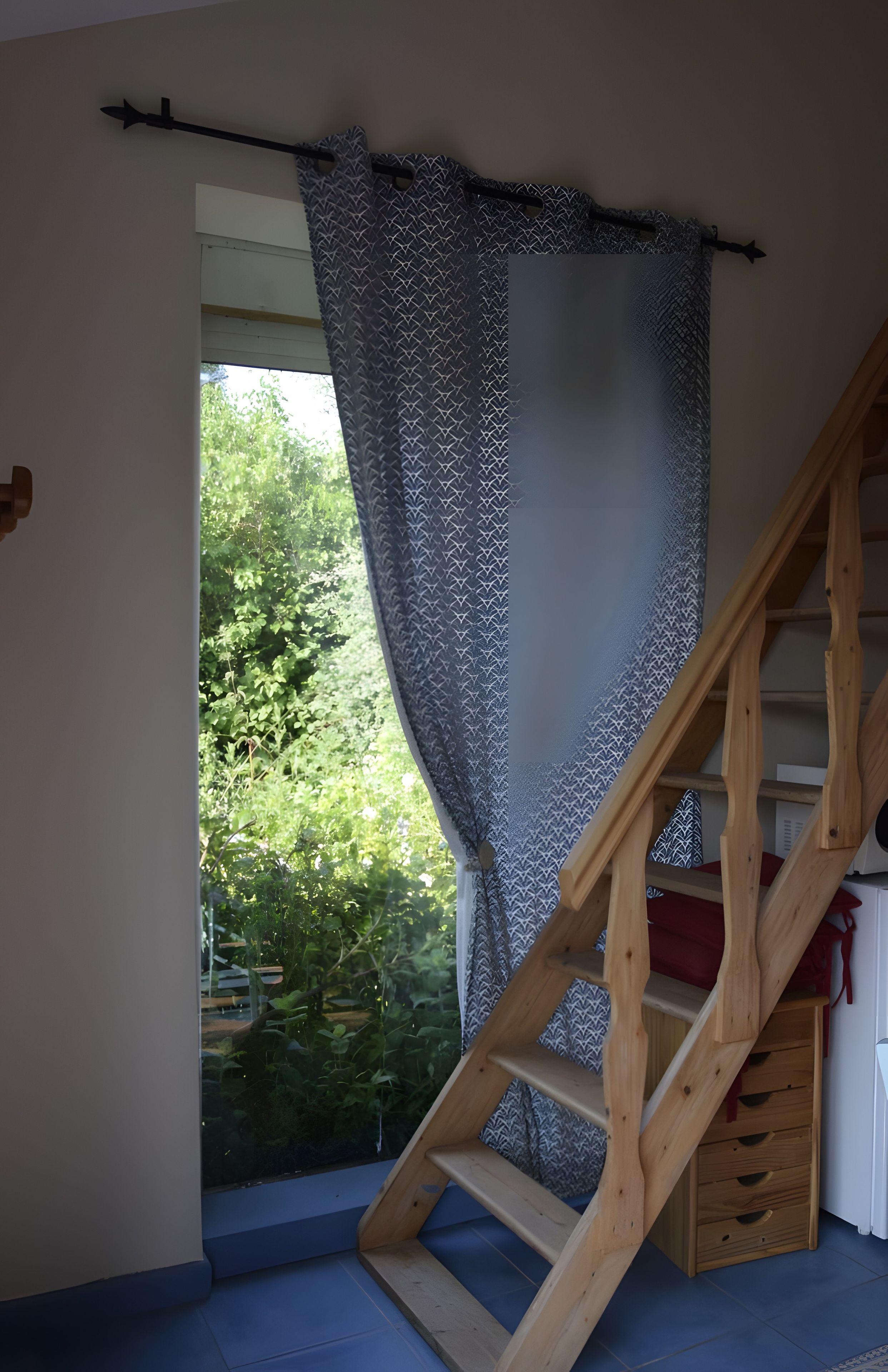 Een kamer met een houten trap, een raam met gordijnen met een patroon, en uitzicht op groen buiten.