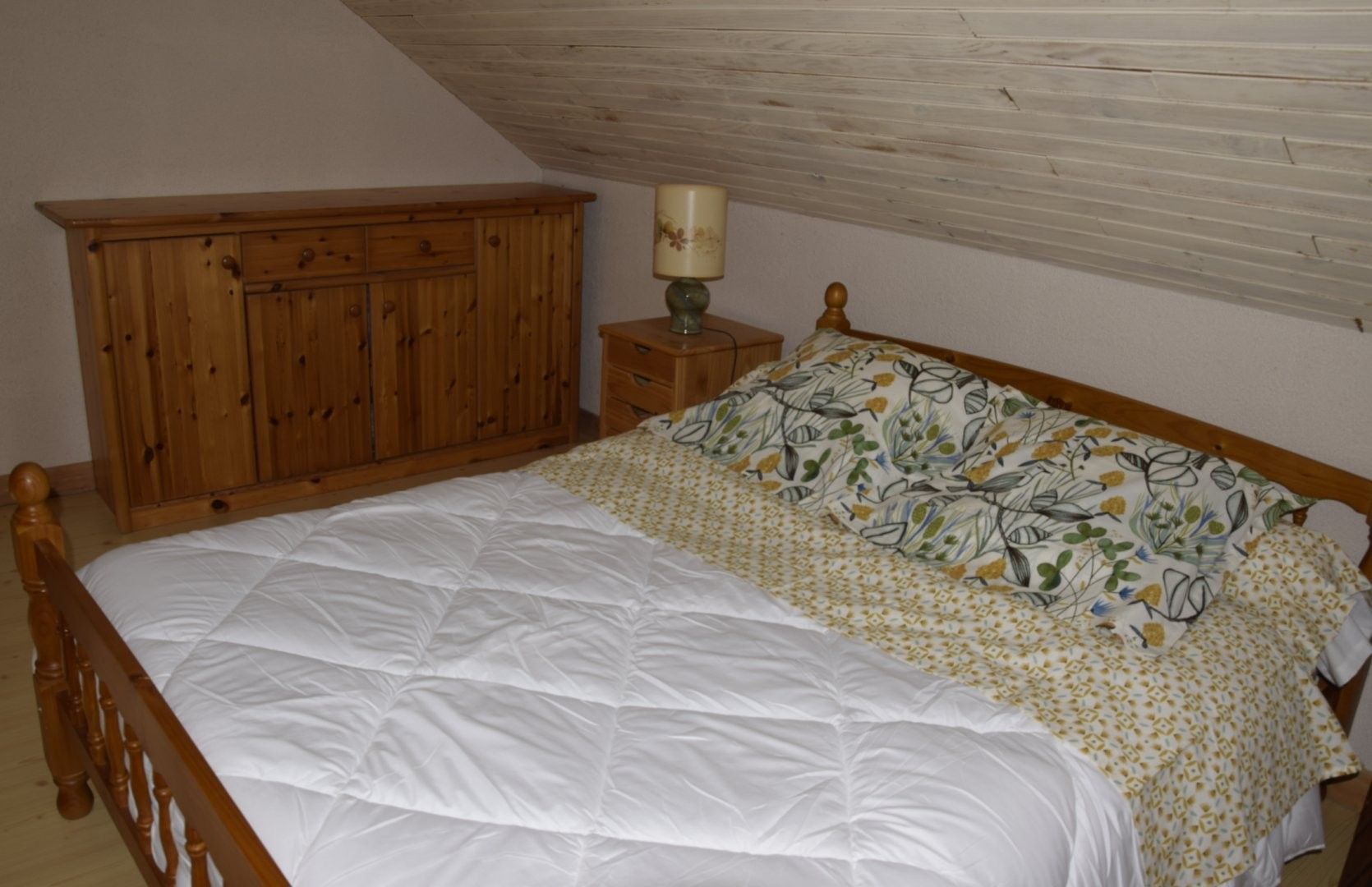 Een slaapkamer met een houten bedframe, een wit dekbed, kussens met een patroon, een houten kledingkast en een bedlampje.