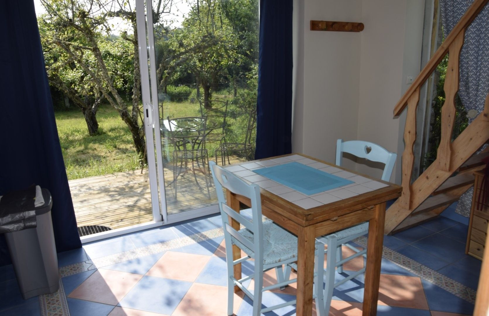 Een kamer met een tafel, stoelen, een houten trap en uitzicht op een tuin door een glazen deur.