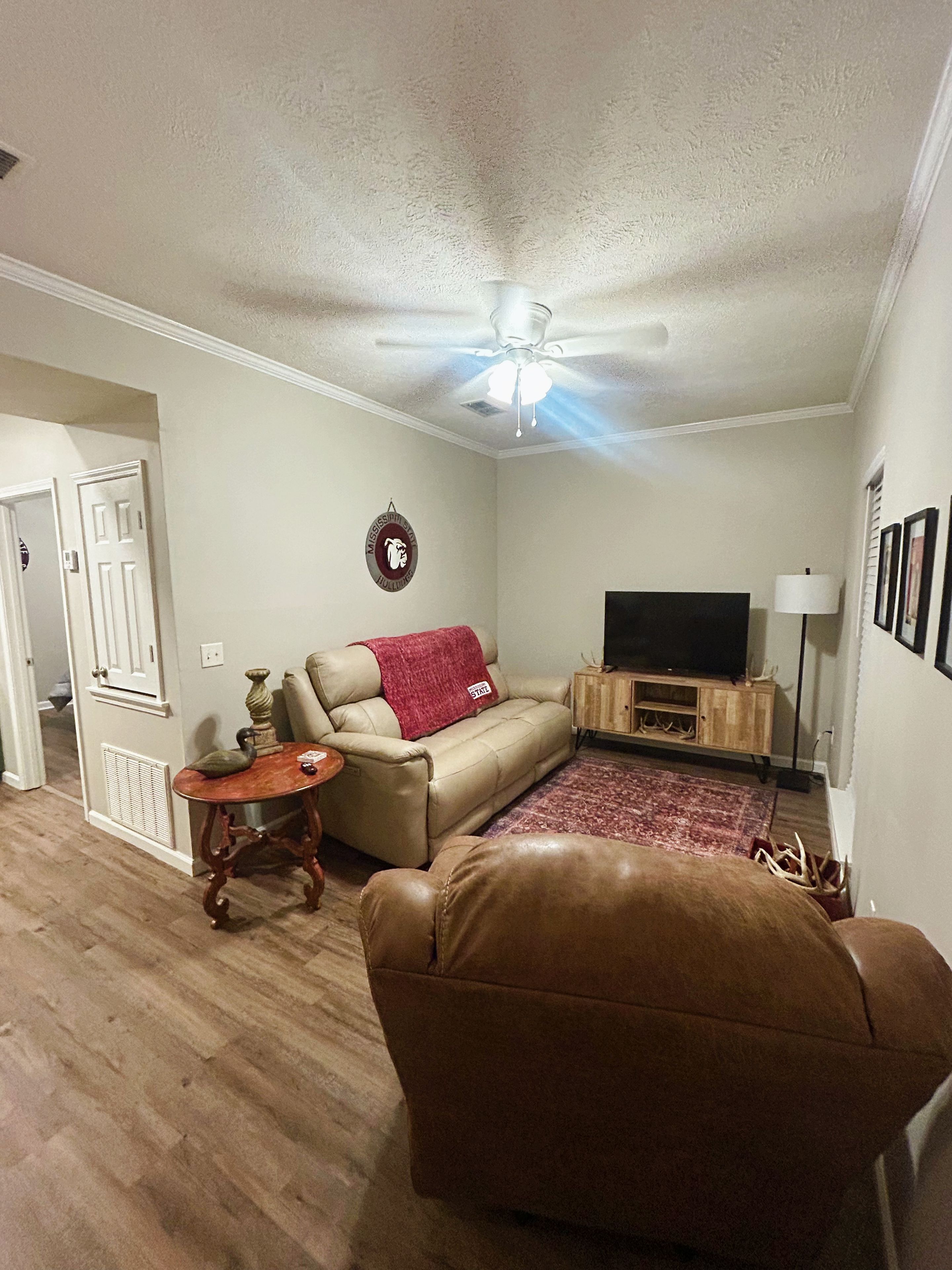 A living room with a sofa, a television, a ceiling fan, and a decorative wall hanging.