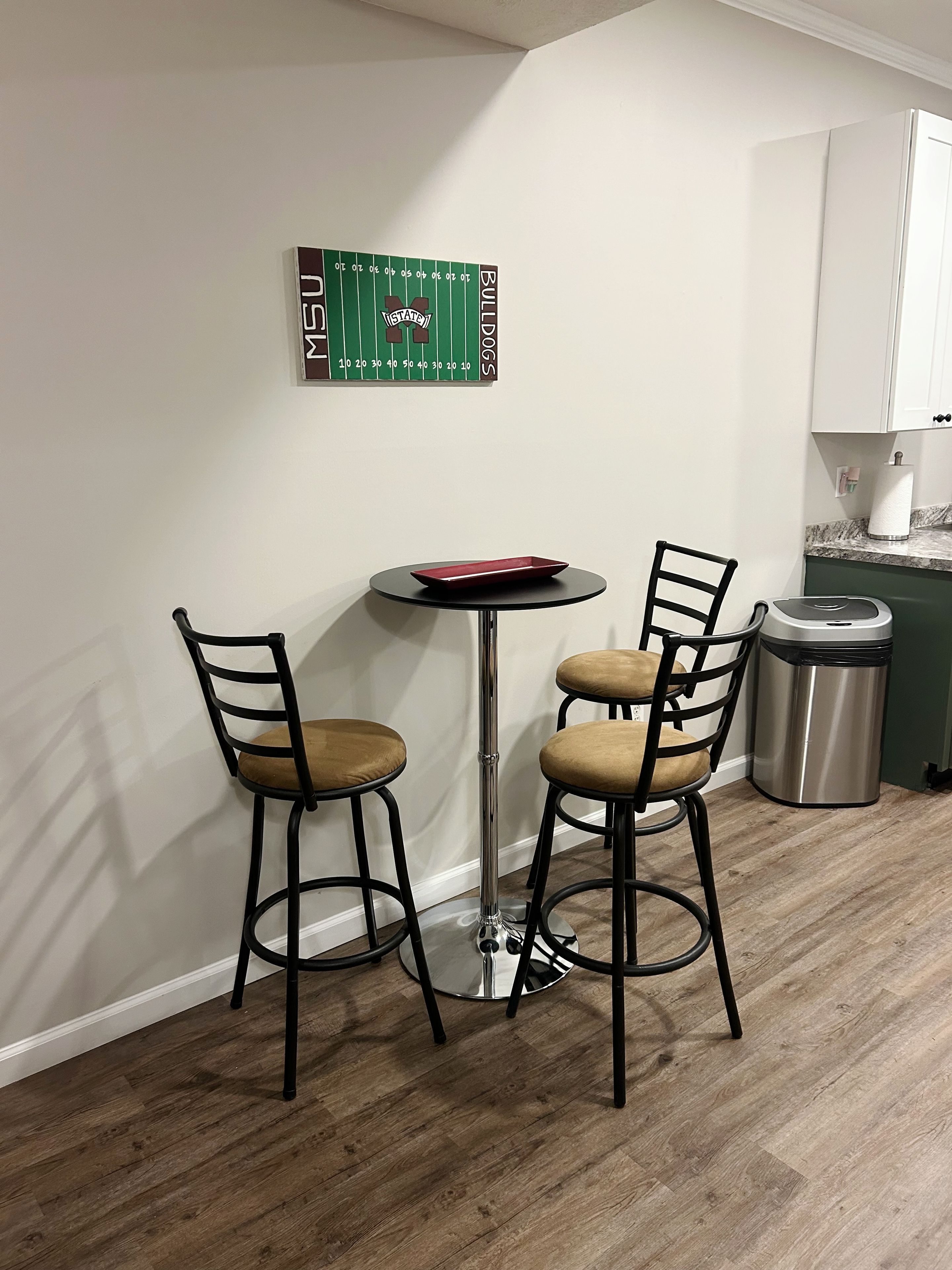 A small dining area with a round table and two chairs, a trash can, and a kitchen counter with cabinets.