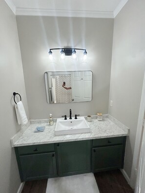 Un baño con lavamanos blanco, armarios verdes, un espejo y una lámpara sobre el espejo.