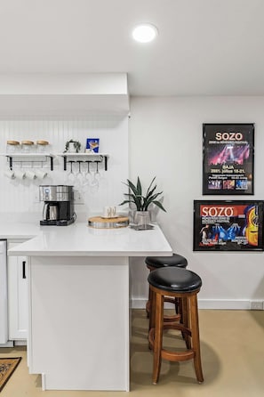 A modern kitchen with a white countertop, hanging mugs, a coffee machine, and a potted plant. There are two stools and a framed poster on the wall. - Cozy Retreat Apartment | Near Downtown Nash (Nashville)