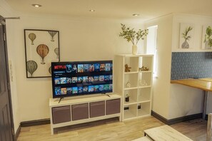 Een woonkamer met een flatscreen televisie waarop een Netflix-interface wordt weergegeven, een wit tv-meubel, een kleine plant en een keukenhoek met een betegelde spatwand.