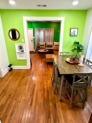 Ein Essbereich mit einem Holztisch und Stühlen, einer grünen Wand und Blick in ein Wohnzimmer mit Sofa und Vorhängen.