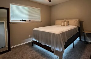 A bedroom with a bed, a window with blinds, a door, and a small table. - Relax & Unwind - Welcoming 2BR Texas Retreat (Waxahachie)