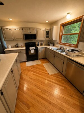 A kitchen with wooden flooring, white cabinetry, a built-in oven, a microwave, a sink, and a window providing a view of greenery outside. - Peaceful Moultonborough Getaway | Sleeps 14 (Moultonborough)