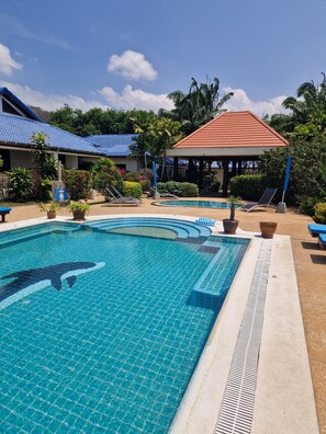 Ein Swimmingpool mit Delfin-Mosaik, umgeben von Liegestühlen, einem Pavillon und tropischer Bepflanzung.