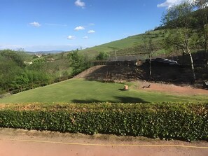 Ein grasbewachsenes Feld mit einer Hecke im Vordergrund, einem Feldweg und einem von Bäumen umgebenen, eingezäunten Bereich.
