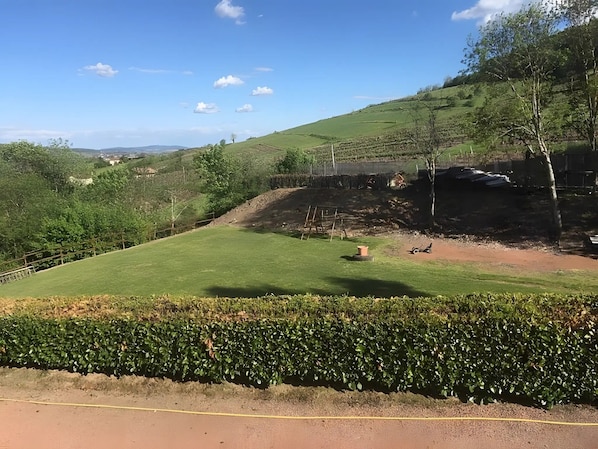 Un prato erboso con un siepe in primo piano, un sentiero sterrato e un'area recintata con alberi.