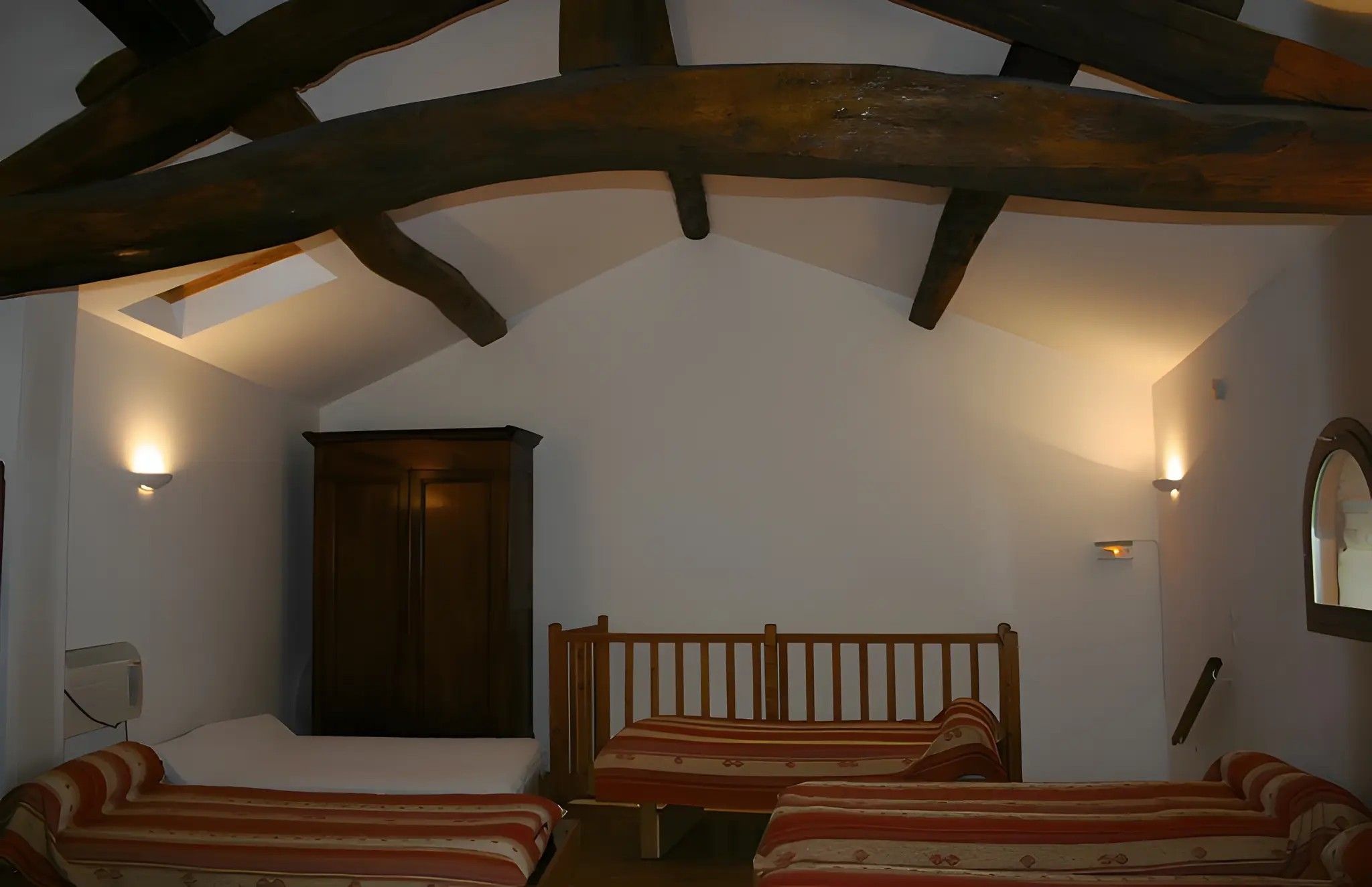 A room with a sloped ceiling, wooden beams, a bed with striped bedding, and wall-mounted lighting.