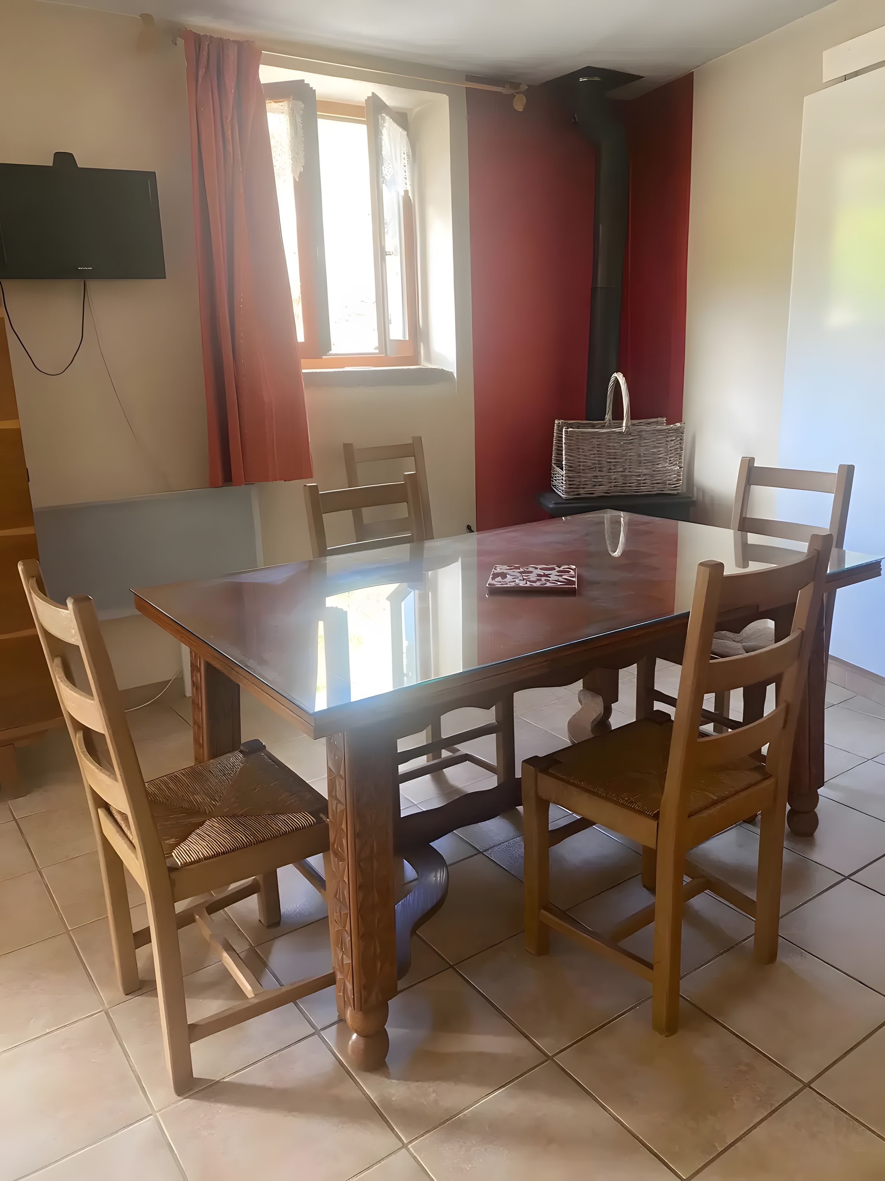 Un coin repas comprenant une table à plateau en verre, des chaises en bois, un panier en osier et un téléviseur fixé au mur.
