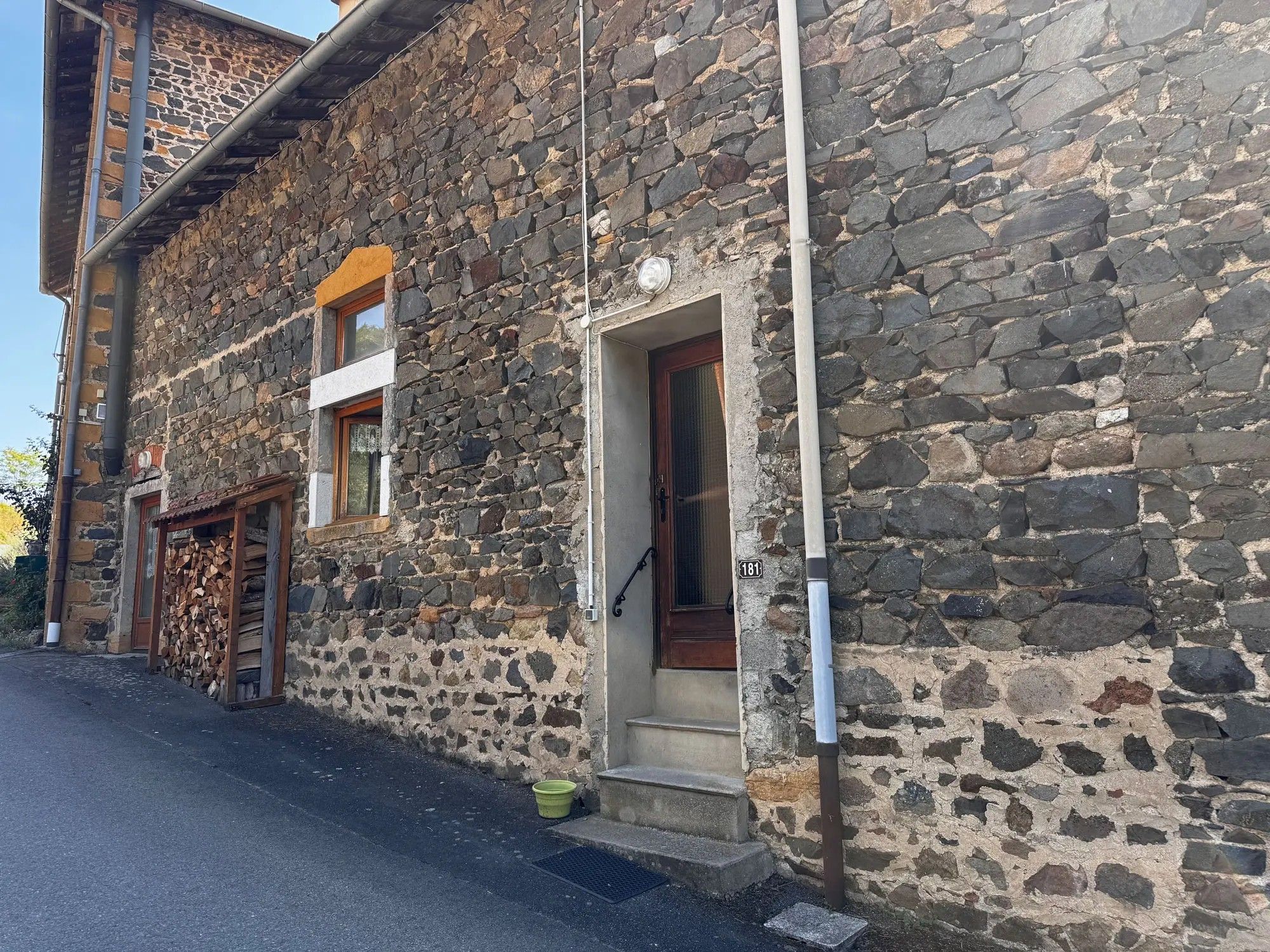 Un edificio de piedra con una puerta de madera y una ventana, un pequeño escalón que lleva a la entrada, y un cubo verde en el suelo.