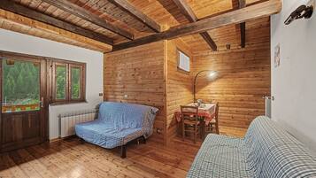 A log cabin interior with a bed, a table with chairs, and a window with a view of trees.