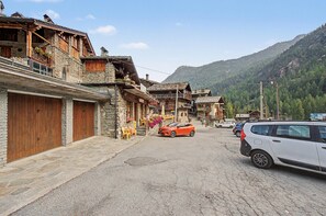 A mountain village street with stone buildings, parked cars, and a view of the mountains.