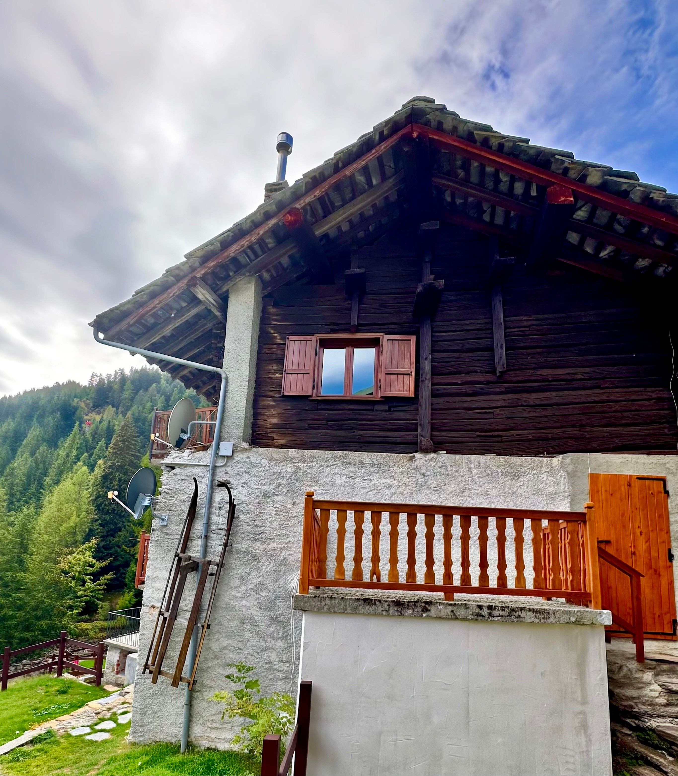 A rustic wooden house with a stone base, a wooden balcony, and a view of a forested mountain.