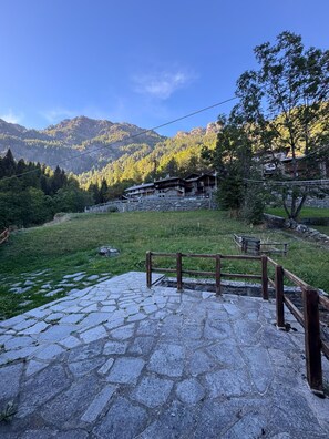Ein steiniger Weg führt zu einem hölzernen Zaun, im Hintergrund eine Berglandschaft und Gebäude.
