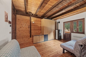 A cabin interior with wooden walls and ceiling, a bed, a sofa, a small kitchenette, and a window with a view of trees.