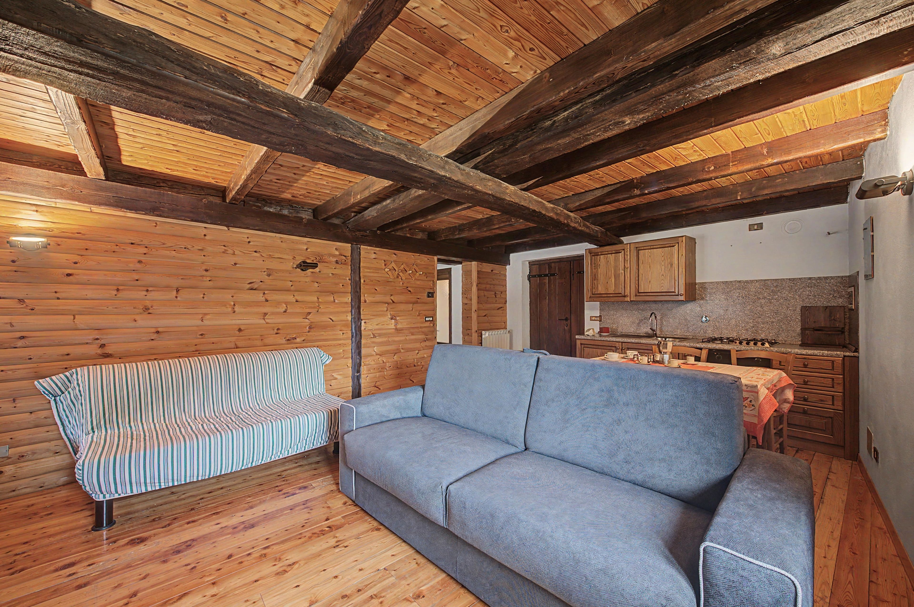 A cozy room with a wooden ceiling, a sofa, a striped chair, and a kitchen area with cabinets and a countertop.