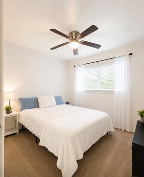 A bedroom with a bed, a ceiling fan, a window with blinds, and a nightstand with a lamp and a plant. - Ashton - Relax & Recharge - Cozy 2BR Retreat (Waxahachie)