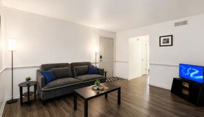 A living room with a sofa, coffee table, TV, and a lamp. - Ashton - Relax & Recharge - Cozy 2BR Retreat (Waxahachie)