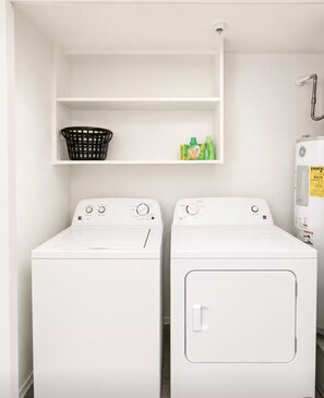 A laundry area with two white washing machines, a black laundry basket, and a shelf with cleaning products. - Ashton - Relax & Recharge - Cozy 2BR Retreat (Waxahachie)