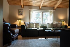 A cozy living room with a green sofa, two armchairs, a coffee table, and a window with curtains. - Lucky Otter: Cozy + Artsy Lakeside Log Cabin (Mount Nebo)
