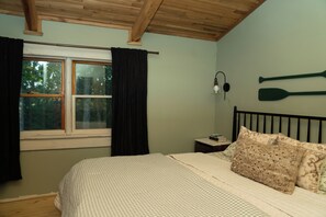 A bedroom with a bed, a window with curtains, a wall-mounted lamp, and two paddles. - Lucky Otter: Cozy + Artsy Lakeside Log Cabin (Mount Nebo)