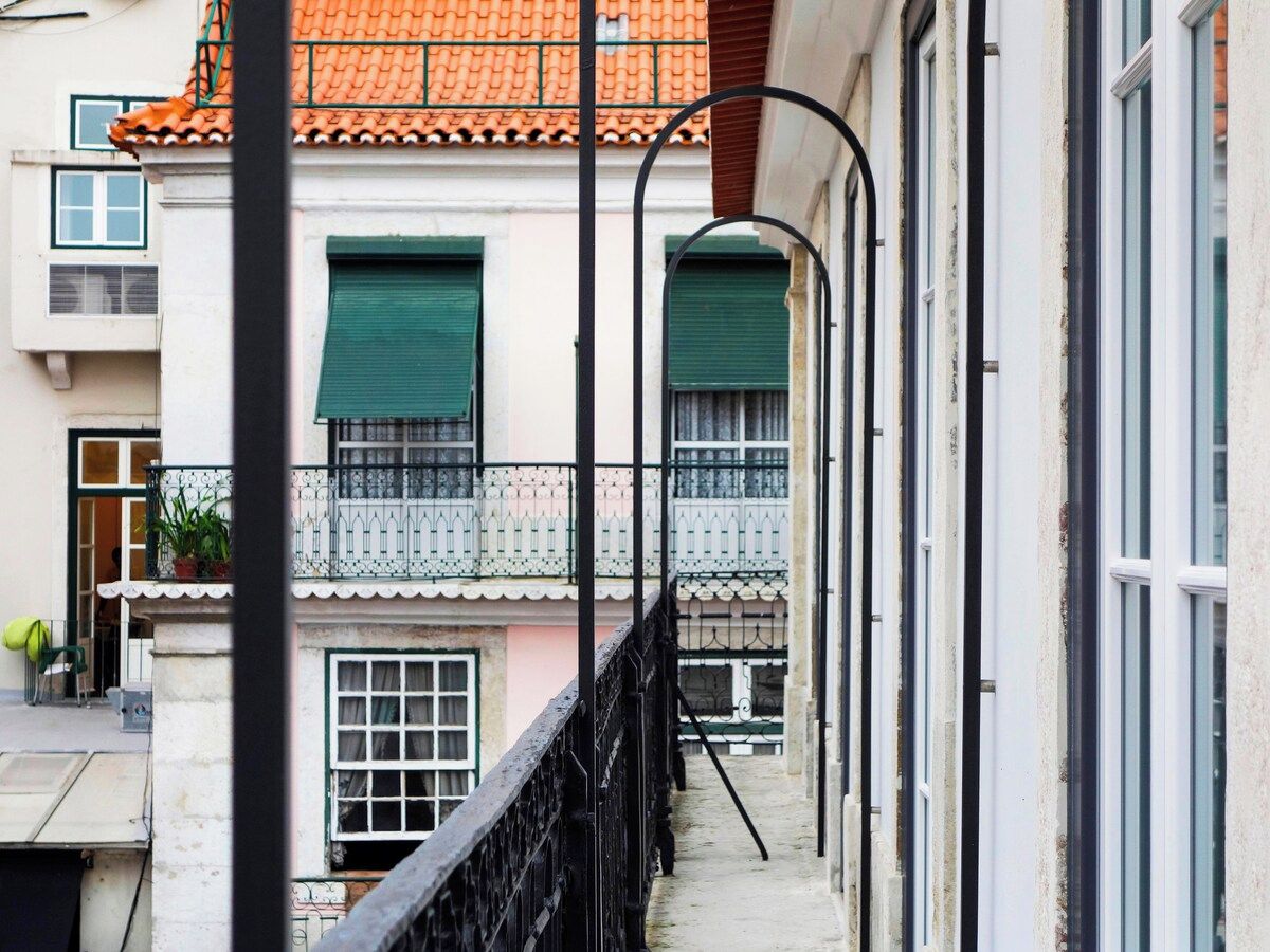 A narrow alleyway with a building featuring a tiled roof, a balcony with a metal railing, and a door with a green shutter.