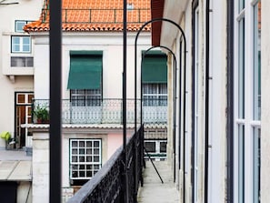 Un estrecho callejón con un edificio que tiene tejado de azulejos, un balcón con baranda de metal y una puerta con contraventanas verdes.