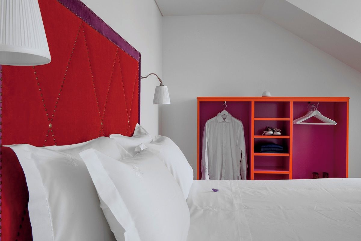A bedroom with a bed, a red headboard, a white lamp, and a pink wardrobe with clothes and shoes.