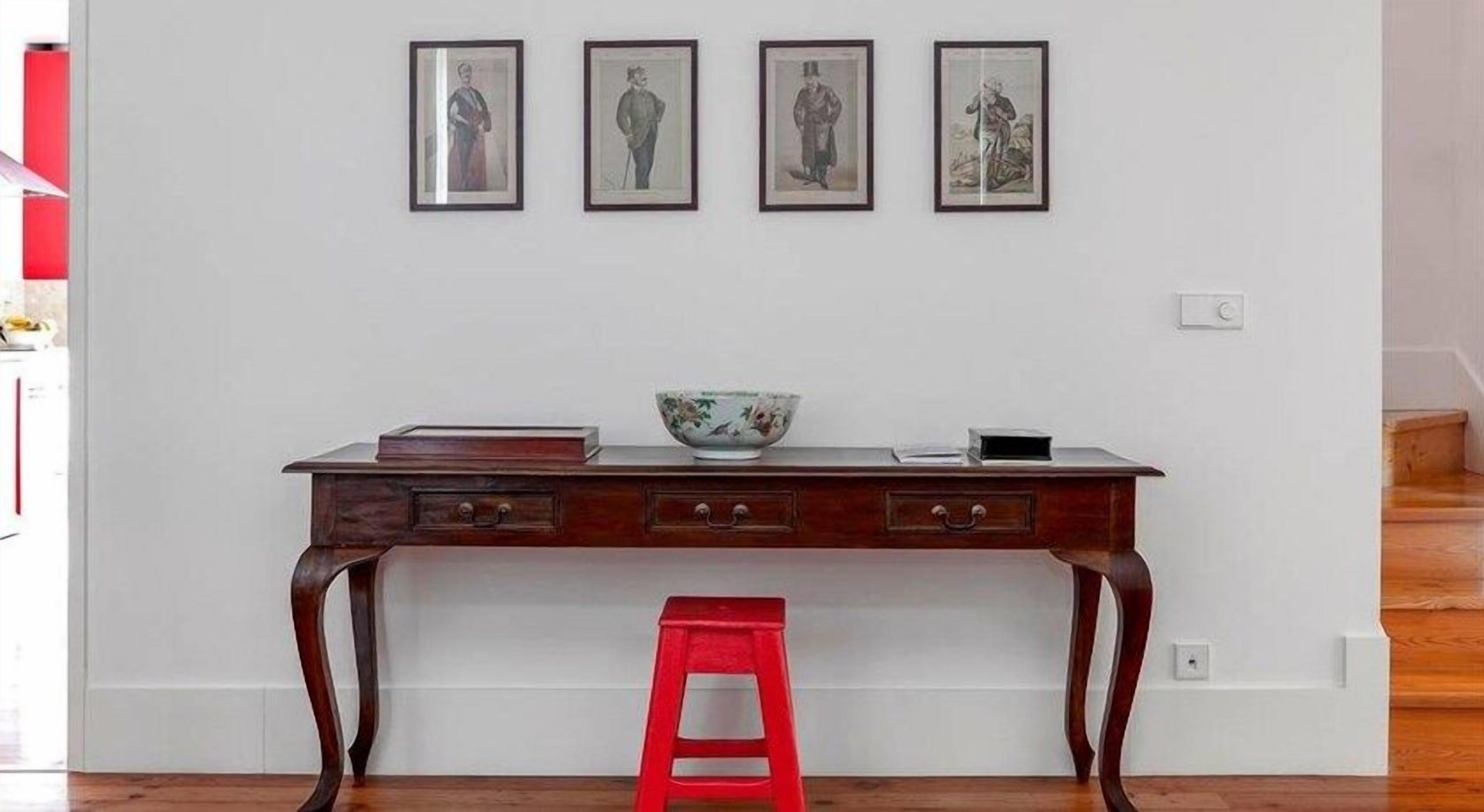 A wooden desk with a red stool in front, a white wall with three framed pictures, and a bowl on the desk.