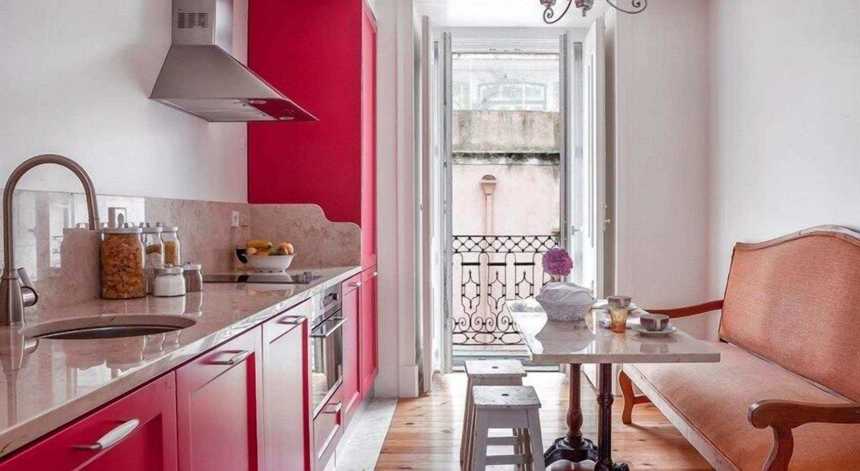 Una cocina moderna con una pared de acento roja, un área de comedor con una mesa blanca y un sofá rosa, y una vista a través de una puerta abierta.