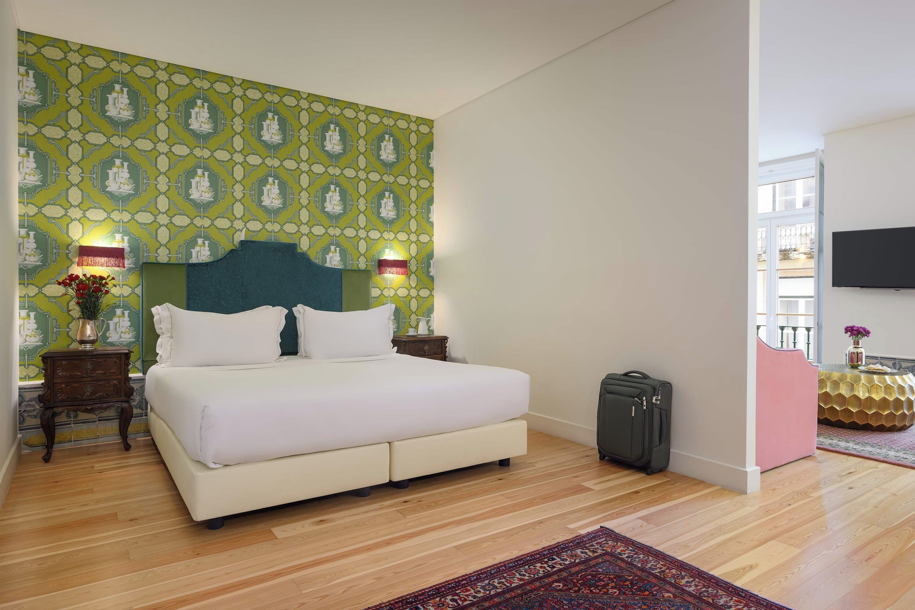 Quarto de hotel com cama grande, piso de madeira, parede verde com padrão, televisão fixada na parede e pequena mesa com um vaso de flores.