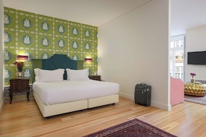 Quarto de hotel com cama grande, piso de madeira, parede verde com padrão, televisão fixada na parede e pequena mesa com um vaso de flores.