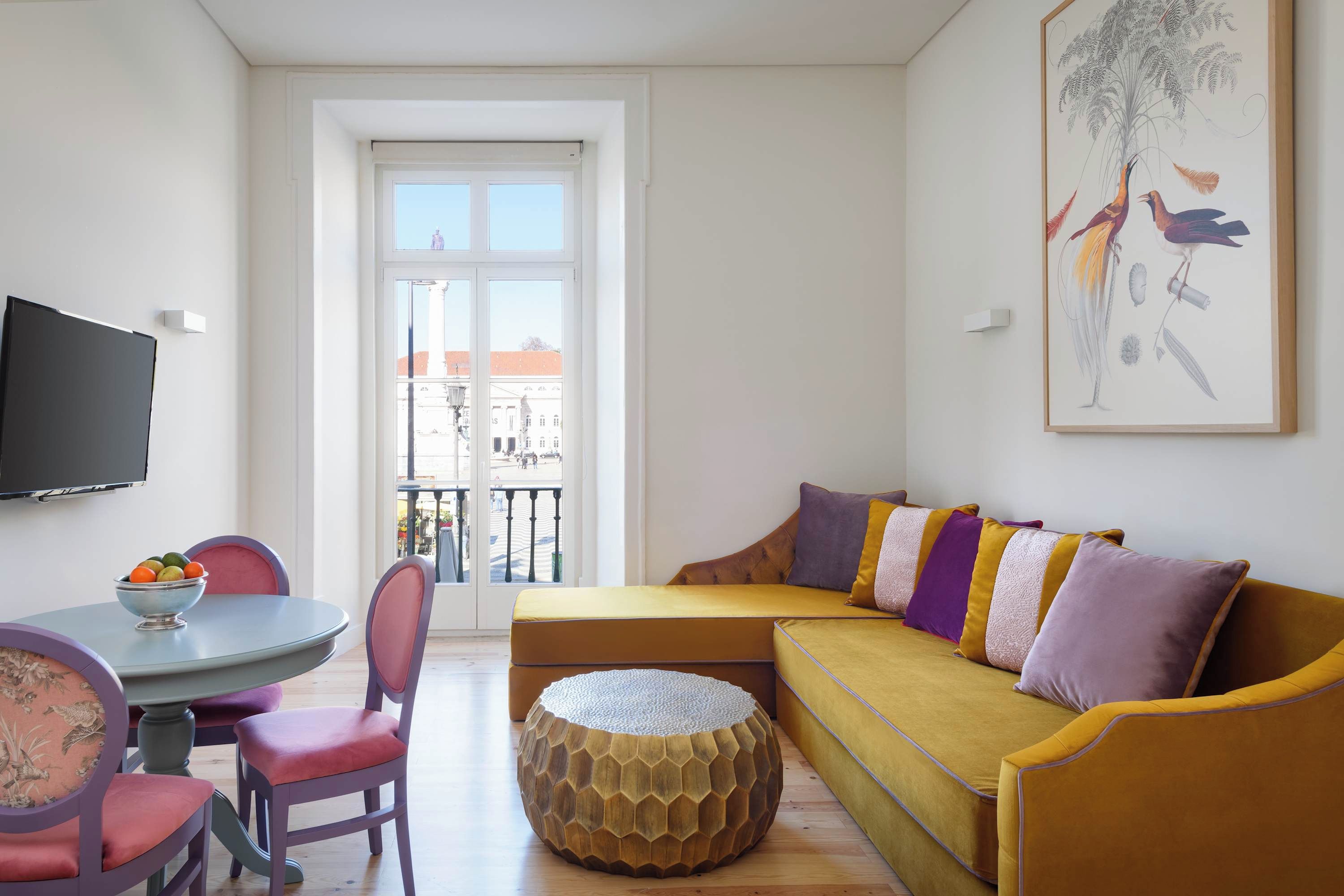 Una sala de estar moderna con un sofá, sillas, un reposapiés de madera redondo, una mesa de comedor con fruta y un ventanal con vistas a edificios.