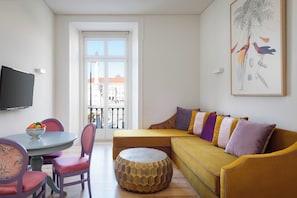 Een moderne woonkamer met een bank, stoelen, een rond houten poef, een eettafel met fruit en een groot raam met uitzicht op gebouwen.