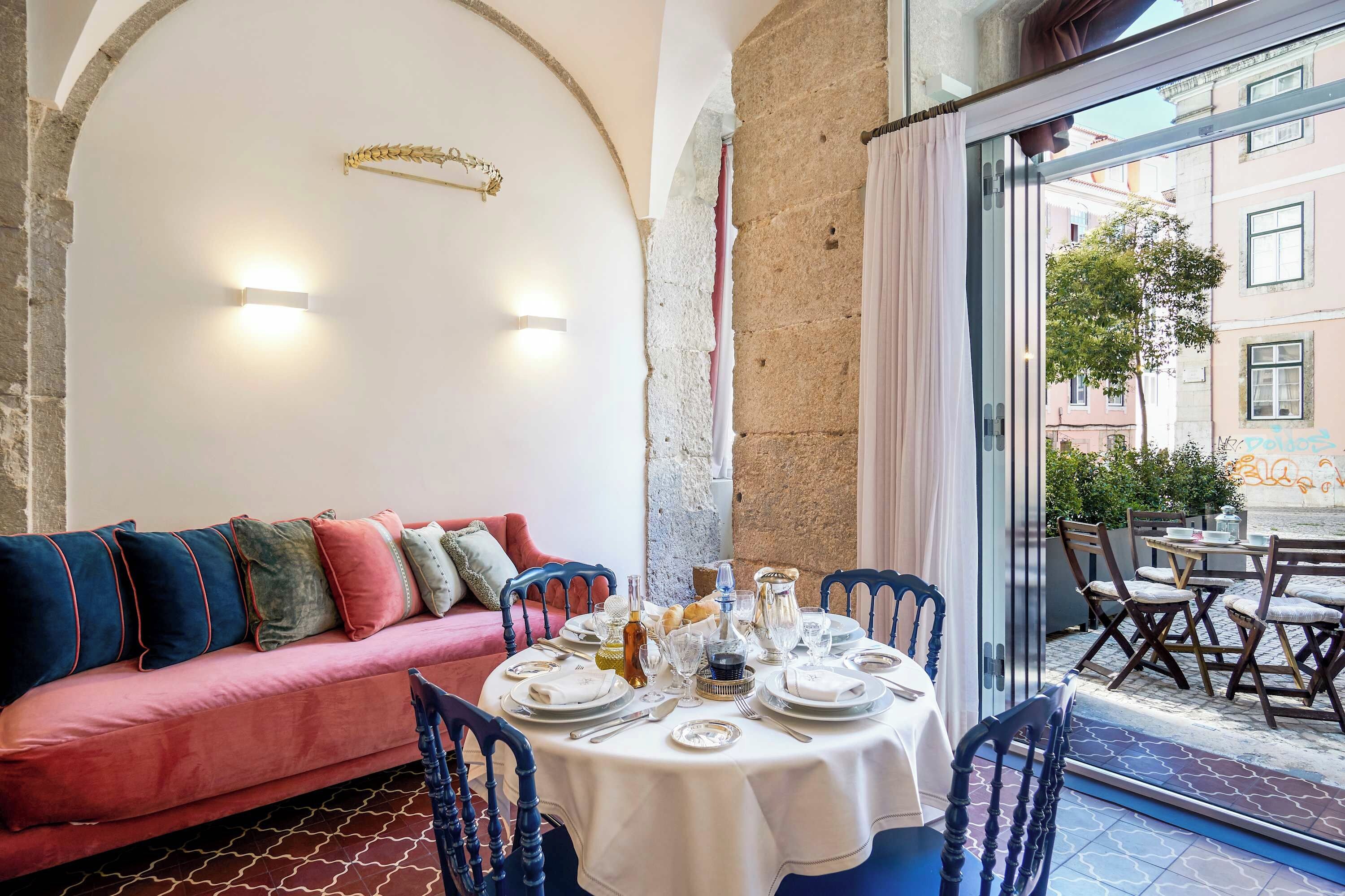 A dining area with a table set for four, a pink sofa with colorful cushions, and a view of the street through a glass door.