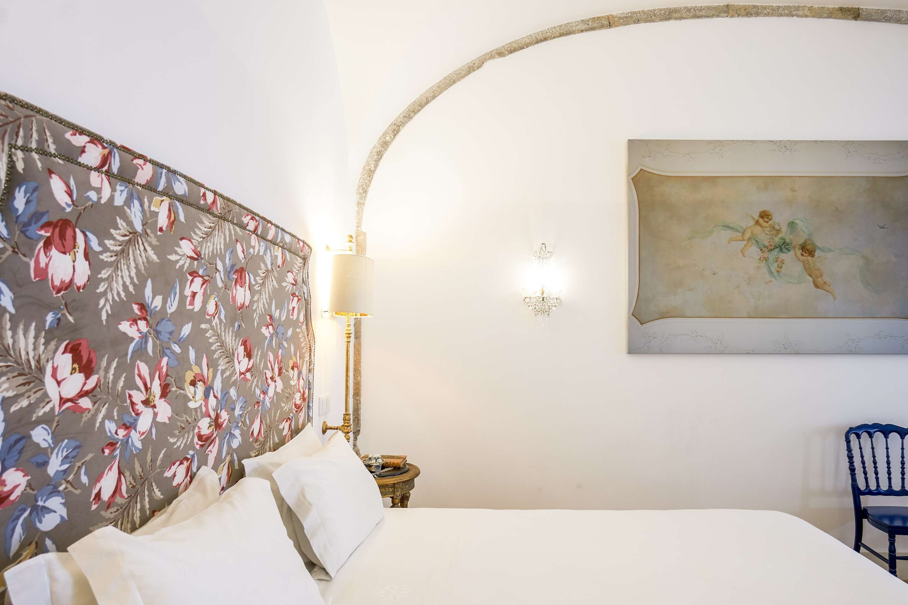 Ein Hotelzimmer mit einem blumengemusterten Kopfteil, einem Bett mit weißen Leinen, einem an der Wand hängenden Gemälde von Cherubs, einer Lampe im klassischen Stil und einem blauen Sessel.