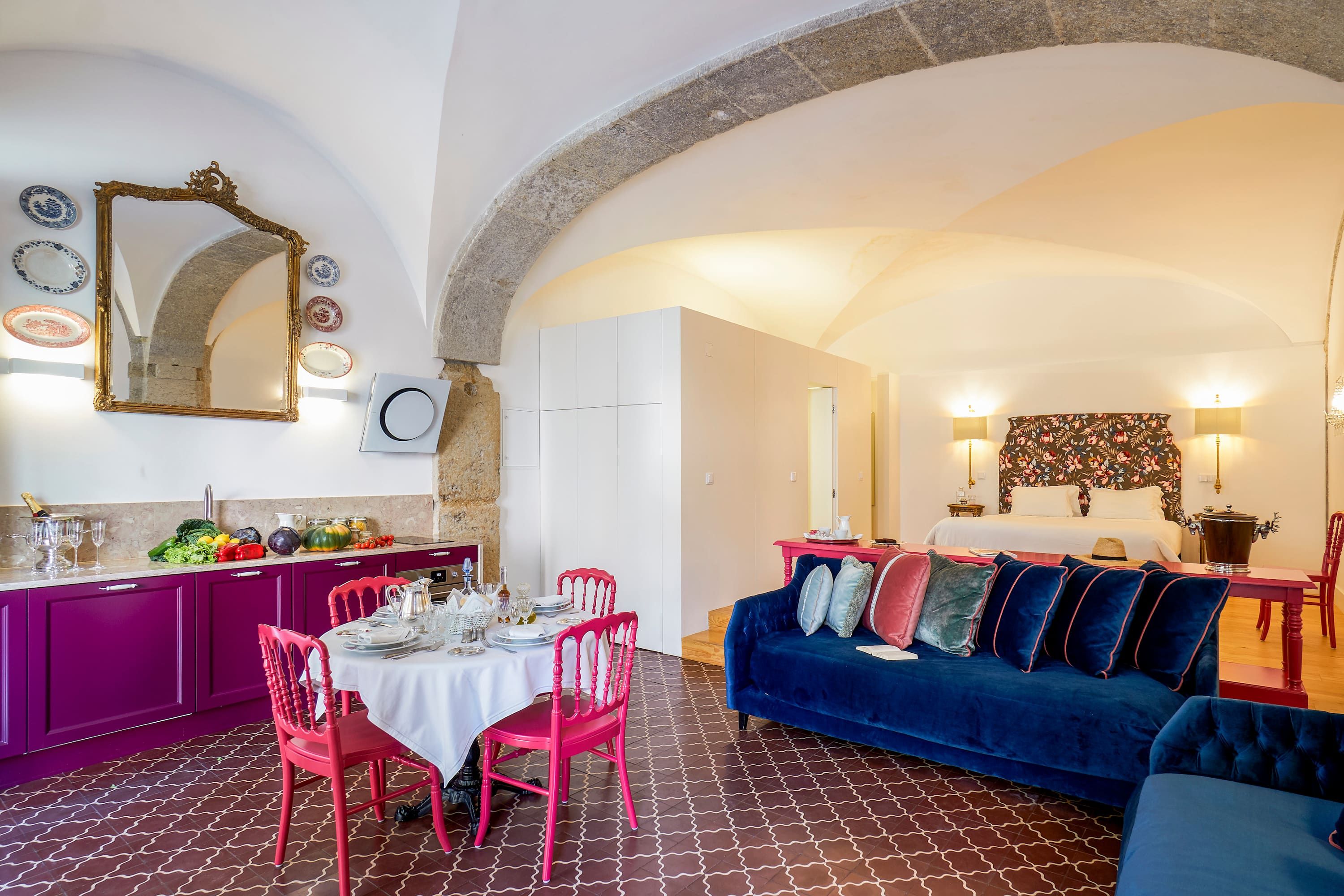 A dining area with a table set for four, a blue sofa, and a kitchen with a stone archway.