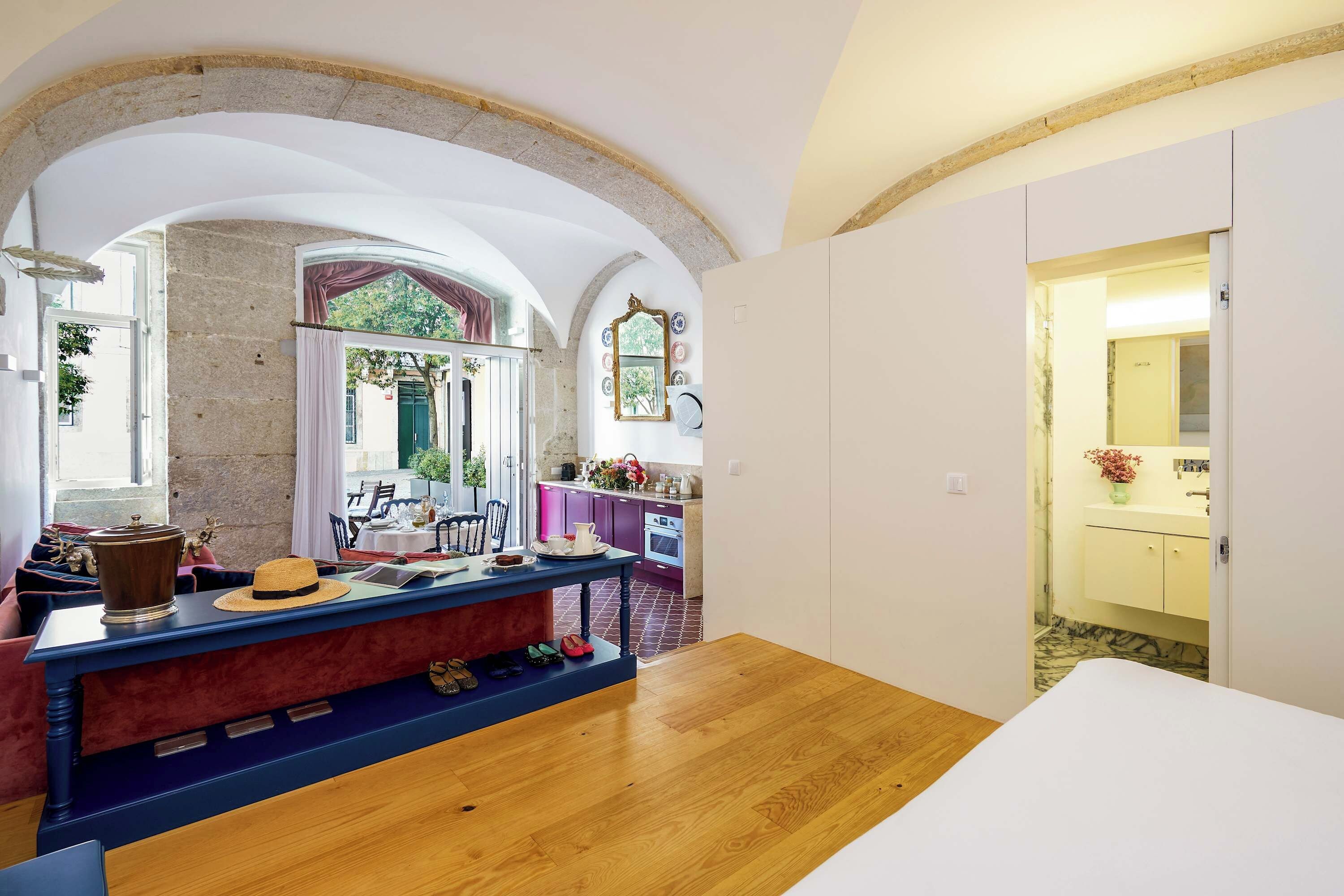 Una habitación con piso de madera, una mesa de comedor con sillas, una zona de cocina con armarios morados y un baño con lavamanos amarillo.
